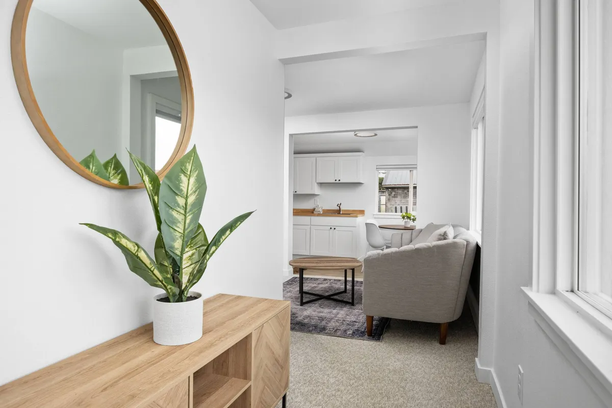 This image centers on a welcoming living area with a round mirror, indoor plant, wooden side table, and a plush sofa. Bathed in morning sunlight, the scene exudes warmth and a relaxed vibe, ideal for unwinding or socializing after beachside adventures. The chic modern furnishings and overall brightness echo the sanctuary theme of the section.