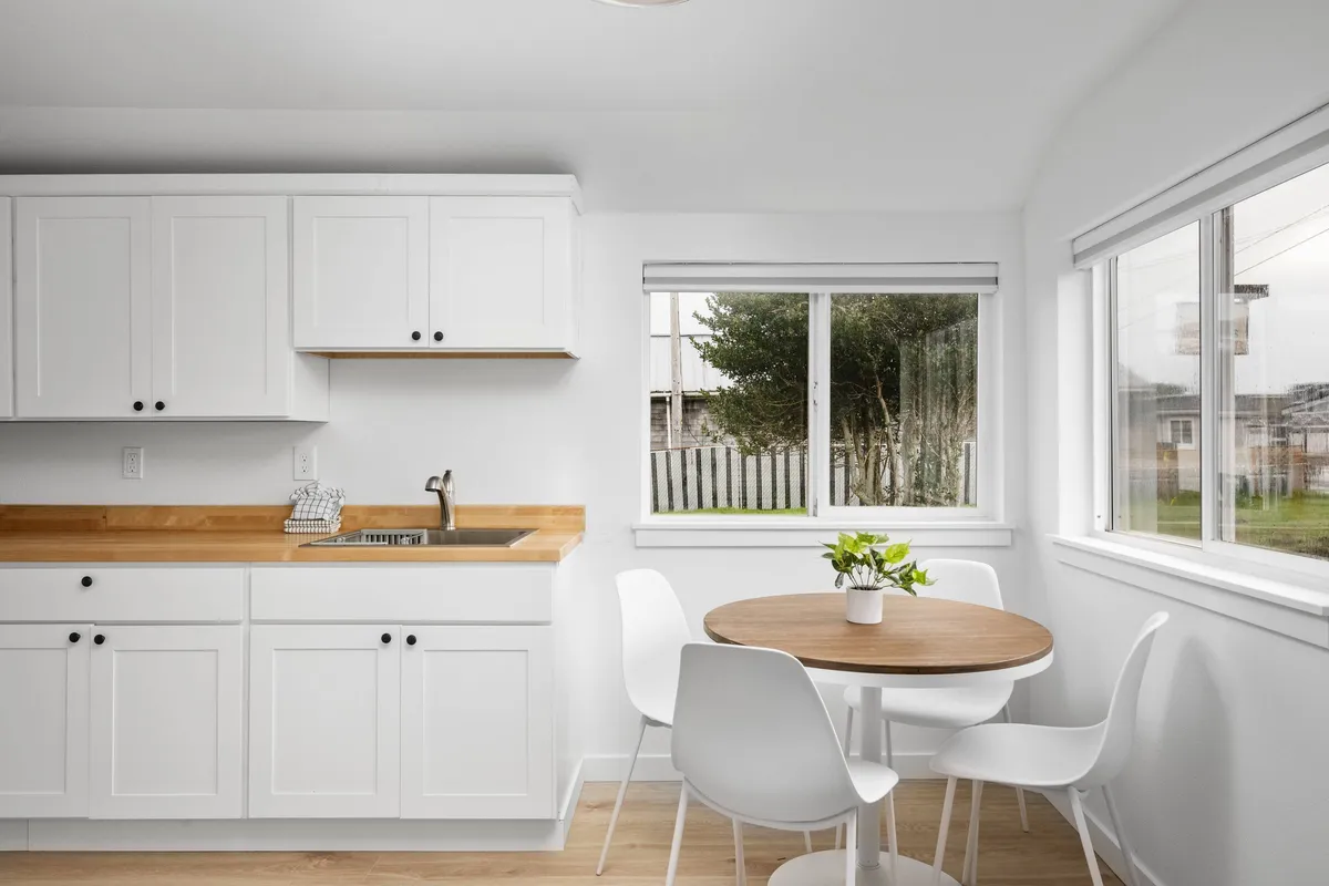 This image captures a bright, welcoming kitchenette with wooden cabinets, a wooden countertop, and a round dining table set near windows that allow natural light to flood the space. The scene exudes a cozy, sociable atmosphere, perfect for either morning brunches or relaxed dinners.