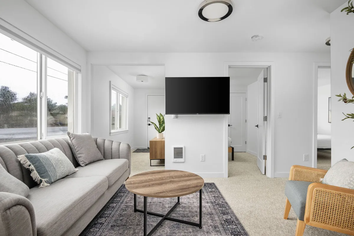 A bright, inviting living room with a cozy sofa and coffee table set near a window, a TV for entertainment, and a touch of greenery from a potted plant. The easy-care flooring is visible, and the space feels warm, comfortable, and practical for both guests and their pets.