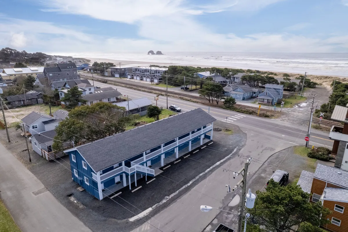 This vibrant exterior shot captures a blue building situated just across from Rockaway Beach, with the ocean visible in the background. The scene also includes other residences, giving a sense of the welcoming community and immediate proximity to the shoreline. The relaxed mood is enhanced by hints of sunset lighting, emphasizing the beachfront atmosphere.